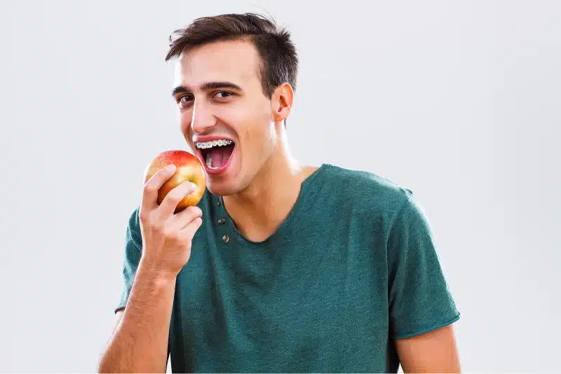Young man with braces smiling and holding an apple, illustrating foods to avoid while wearing braces for better oral health.